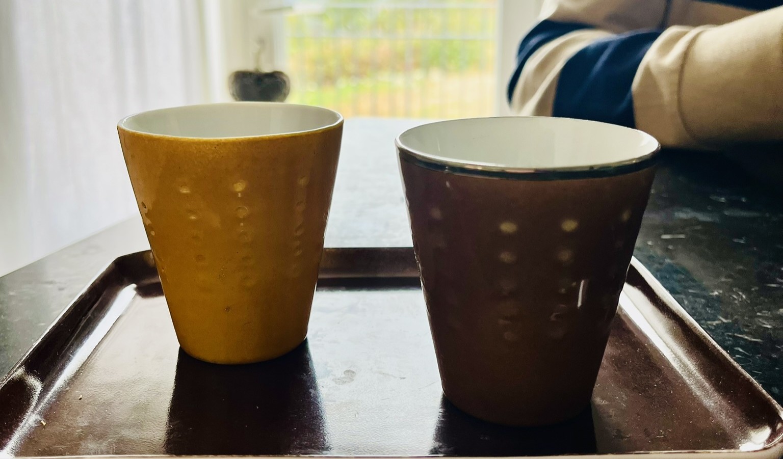 Deux tasses, l'une jaune et l'autre brune, posées côte à côte sur un plateau devant une porte-fenêtre