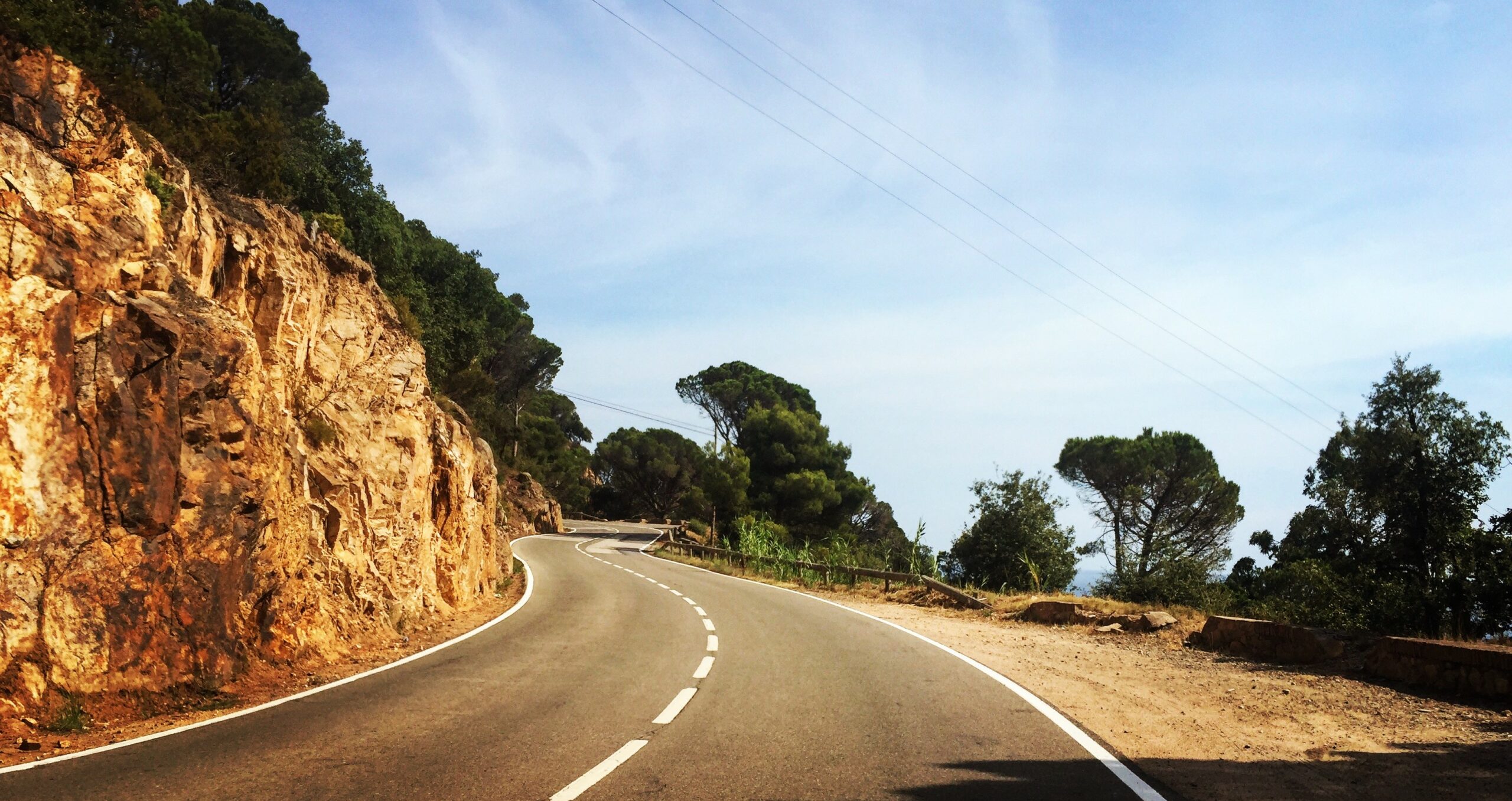 Route de corniche sinueuse longeant une paroi rocheuse orangée, sous un ciel bleu